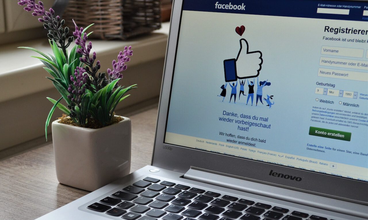 Βλέπω ένα λάπτοπ που εμφανίζει τη σελίδα εγγραφής του Facebook στα γερμανικά. Δίπλα στον υπολογιστή υπάρχει ένα λευκό γλαστράκι με λιλά λουλούδια. Το λάπτοπ είναι της μάρκας Lenovo